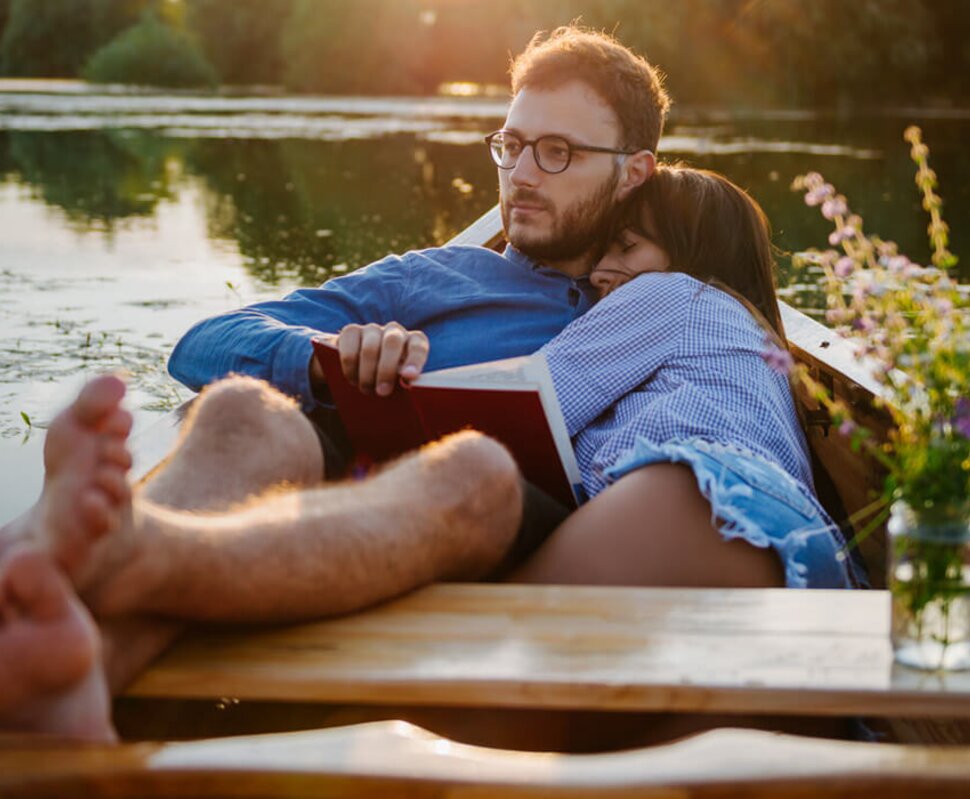 Ein Paar entspannt in einem Boot am Wasser, sie lehnt an ihn, während er ein Buch liest. Sanftes Licht der Abendsonne.