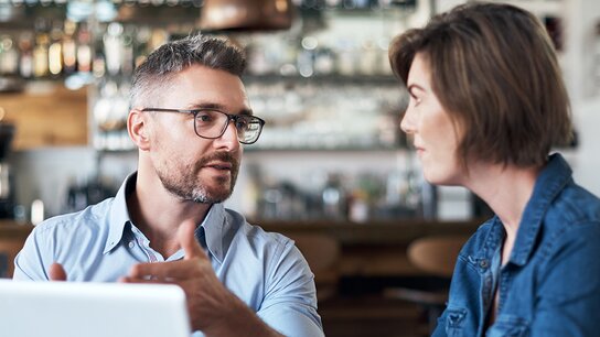 Ein Mann und eine Frau unterhalten sich angeregt in einem Café, im Hintergrund ist eine Bar mit verschiedenen Getränken zu sehen.