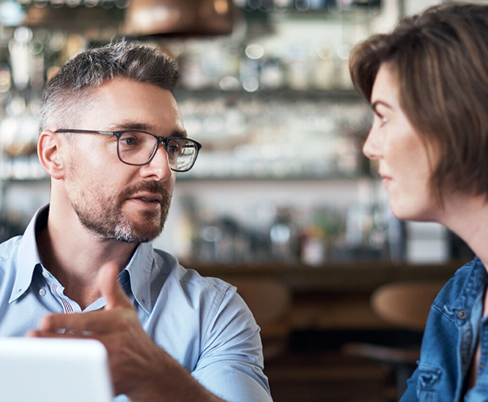 Ein Mann und eine Frau unterhalten sich angeregt in einem Café, im Hintergrund ist eine Bar mit verschiedenen Getränken zu sehen.