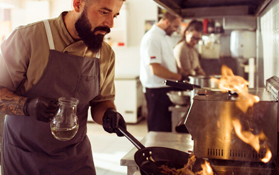 Koch in einem Restaurant bereitet ein Gericht zu, während Flammen aus der Pfanne schlagen und andere Köche im Hintergrund arbeiten.