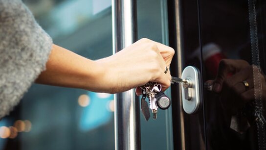 Eine Hand steckt einen Schlüssel ins Schloss einer Glastür, bereit zum Öffnen.