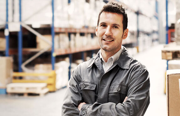 Mann in Arbeitskleidung lächelt in einem Lager mit Regalen voller Waren im Hintergrund.