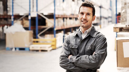 Mann in Arbeitskleidung lächelt in einem Lager mit Regalen voller Kartons im Hintergrund.