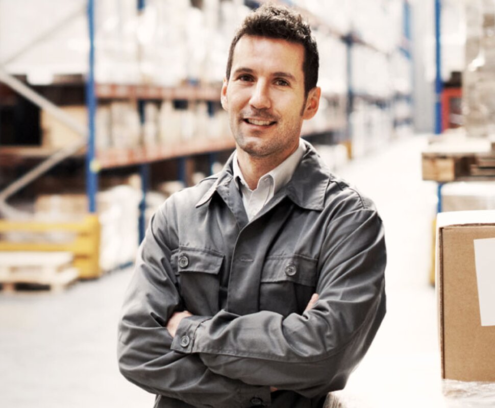 Mann in Arbeitskleidung lächelt in einem Lager mit Regalen voller Kartons im Hintergrund.