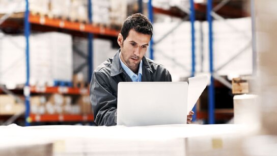Mann in Lagerumgebung arbeitet am Laptop und überprüft Papiere auf einem Tisch. Regale mit Ware im Hintergrund.