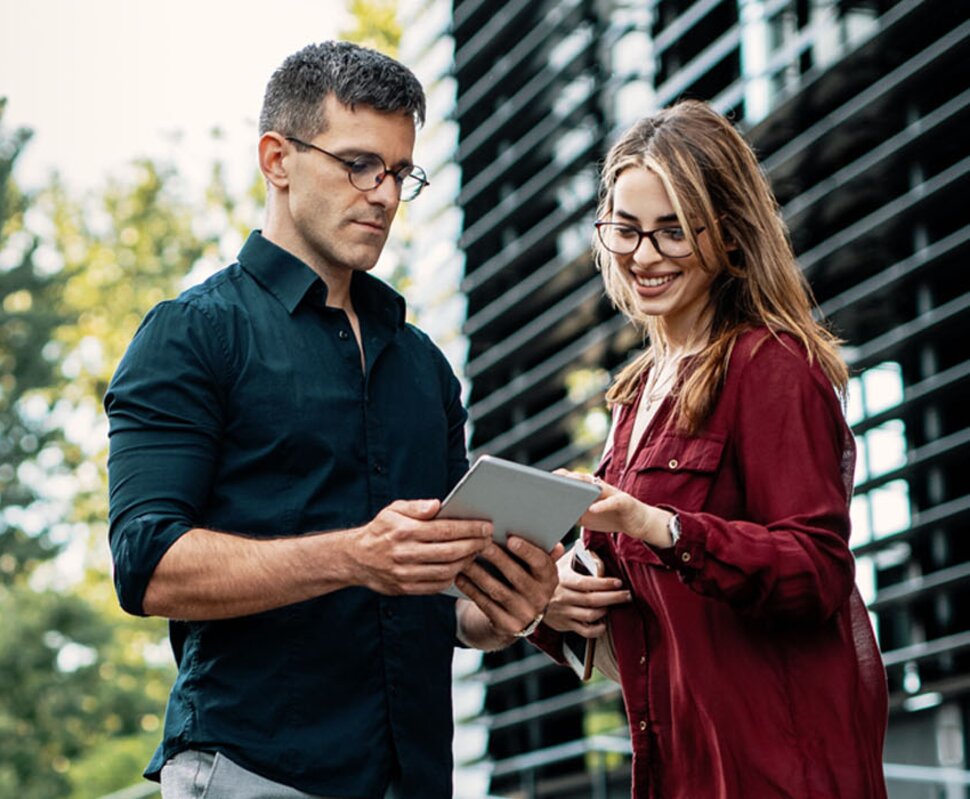 Ein Mann und eine Frau stehen im Freien und schauen gemeinsam auf ein Tablet, umgeben von Pflanzen und modernen Gebäuden.
