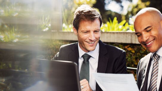 Zwei Männer in Anzügen sitzen lächelnd am Tisch, schauen auf Dokumente und arbeiten gemeinsam an einem Laptop im Freien.