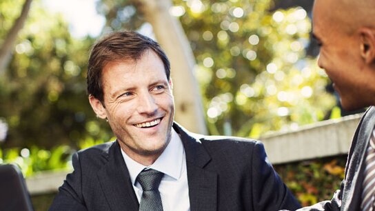 Zwei Männer im Freien unterhalten sich entspannt, während einer von ihnen lächelt. Grüne Pflanzen im Hintergrund.