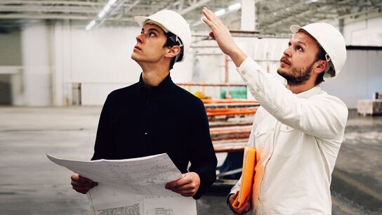 Zwei Bauarbeiter in Schutzhelmen betrachten Baupläne und diskutieren in einem großen, leeren Industriegebäude.