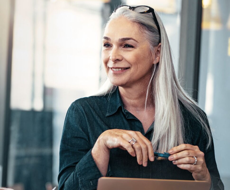 Ältere Frau mit langen grauen Haaren, lächelt und hält einen Stift, während ein Mann im Hintergrund ihr zusieht.