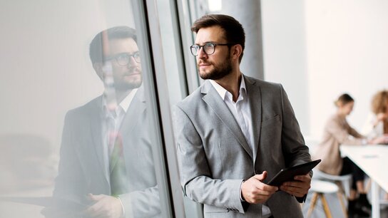 Mann in Anzug hält ein Tablet, schaut nachdenklich aus einem Fenster; im Hintergrund sind weitere Personen bei der Arbeit.