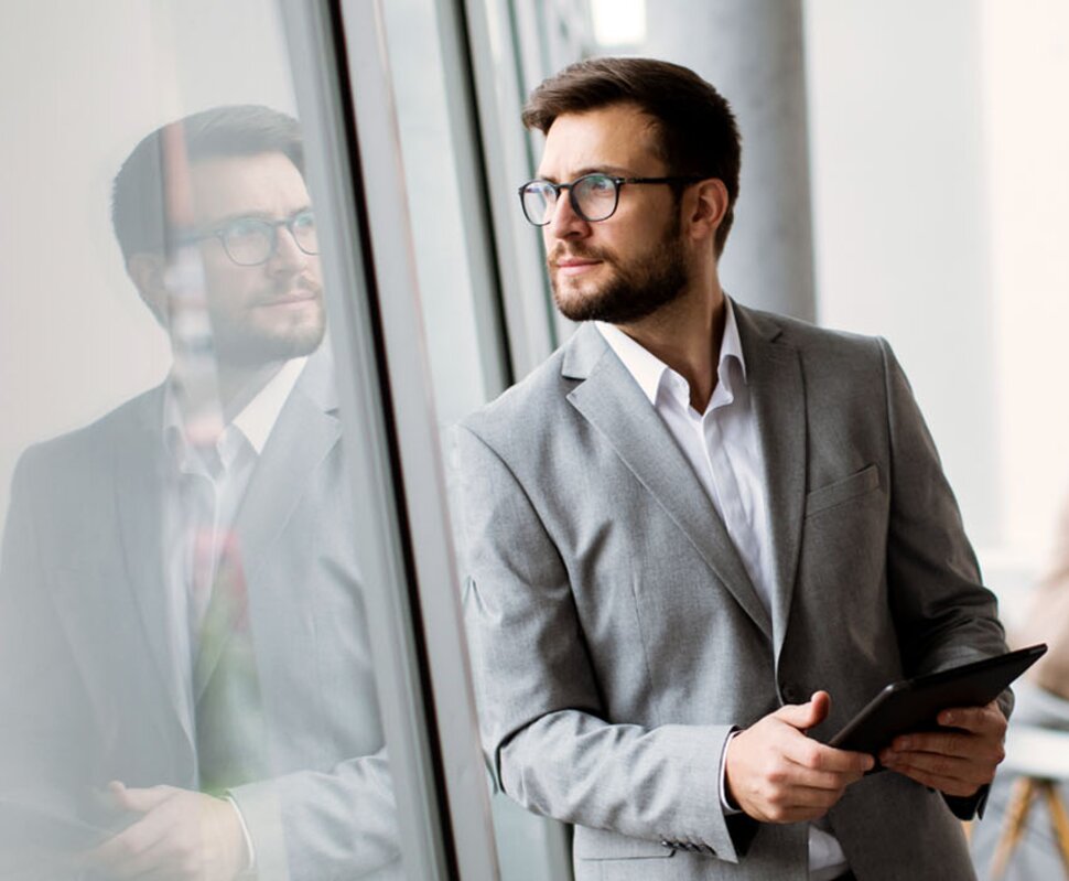 Mann in Anzug hält ein Tablet, schaut nachdenklich aus einem Fenster; im Hintergrund sind weitere Personen bei der Arbeit.