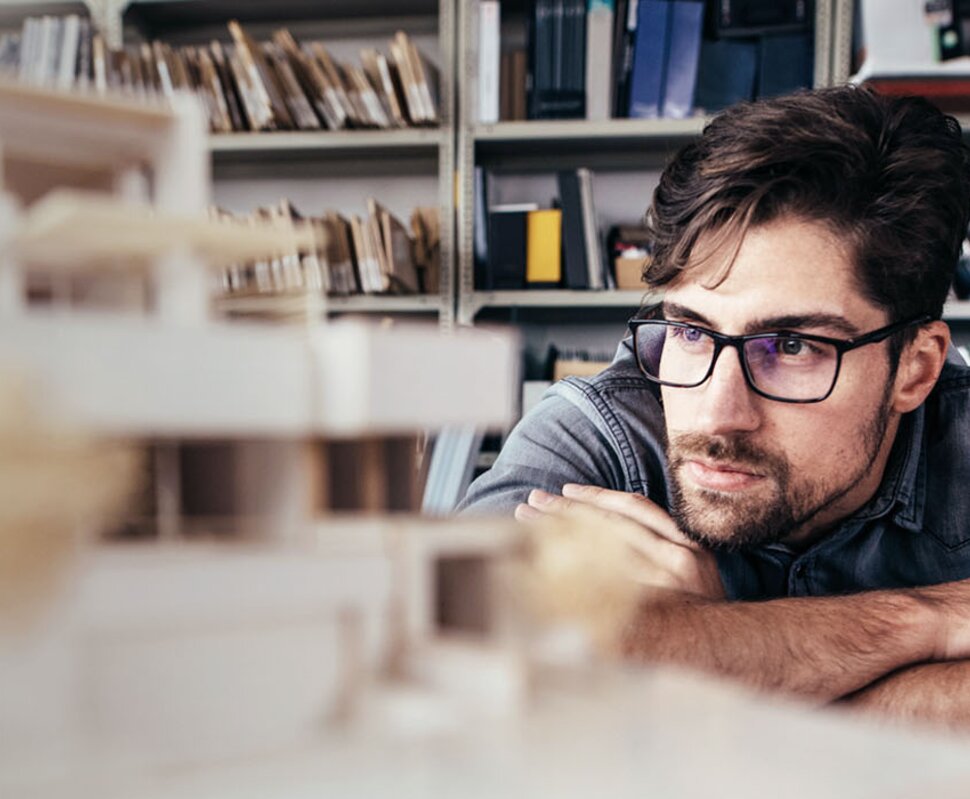 Junger Architekt betrachtet konzentriert ein Modell eines Gebäudes in einem Büro mit Regalen im Hintergrund.