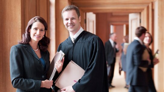 Zwei Menschen, eine Anwältin und ein Richter, posieren lächelnd in einem Gerichtssaal mit Akten in der Hand.