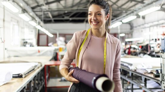Lächelnde Frau in einem Nähatelier hält einen Stoffrollen und ein Maßband in der Hand. Helle, freundliche Arbeitsumgebung.