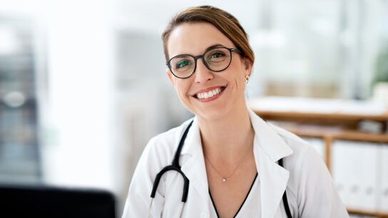Lächelnde Ärztin mit Brille in weißem Kittel, sitzend in einem modernen Behandlungsraum.