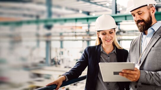 Zwei Fachleute mit Schutzhelmen betrachten gemeinsam ein Tablet in einer industriellen Umgebung.