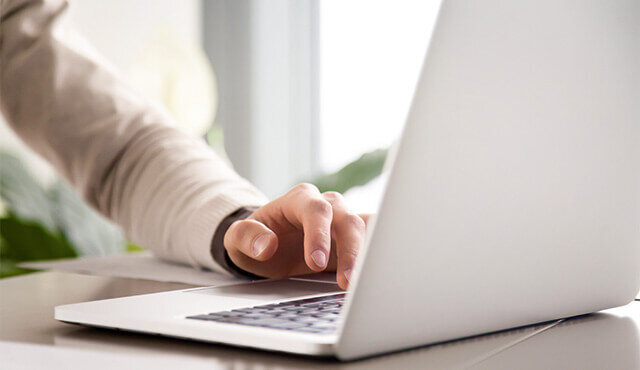 Eine Hand tippt auf der Tastatur eines offenen Laptops, im Hintergrund sind Licht und Pflanzen sichtbar.