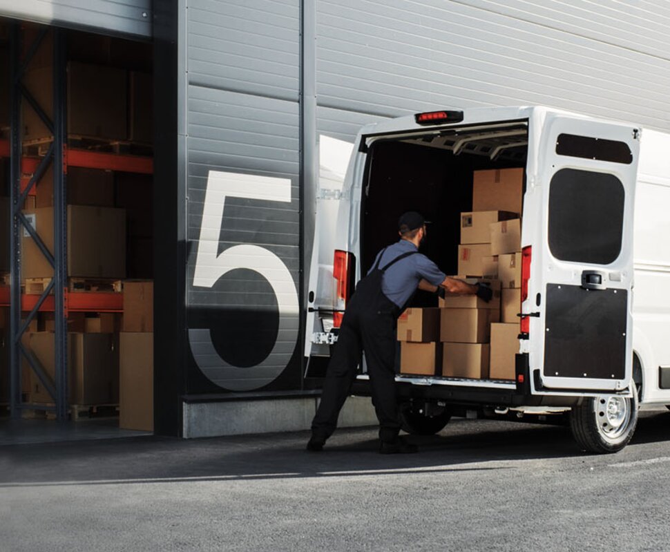 Lieferwagen wird entladen, während ein Arbeiter Kartons aus dem Rückraum herausnimmt, vor einem Lagerhaus mit Regalen.