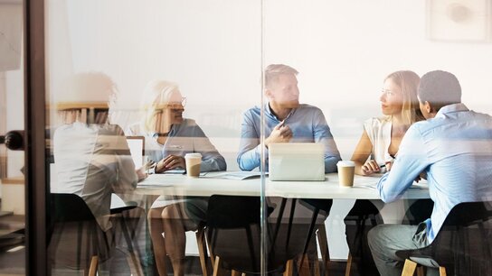 Sechs Personen in einem Konferenzraum, die an einem Tisch sitzen und ein Gespräch führen, mit Laptops und Kaffeetassen.