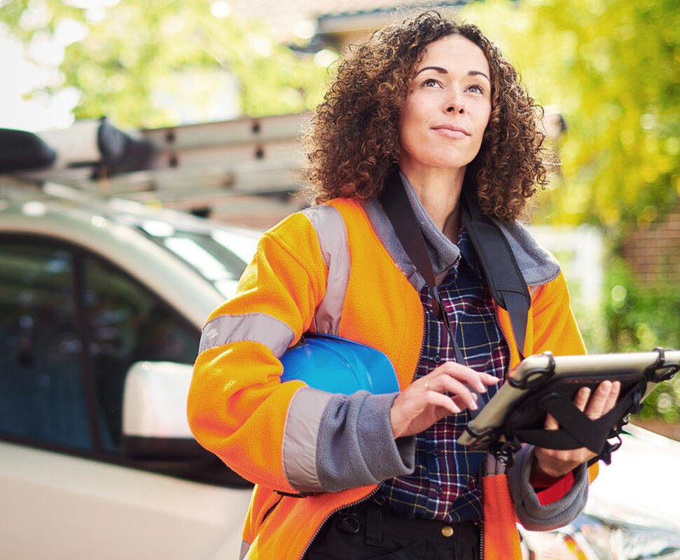 Frau in Sicherheitskleidung mit Helm, die auf einem Tablet arbeitet, steht neben einem Lieferwagen in einer grünen Umgebung.