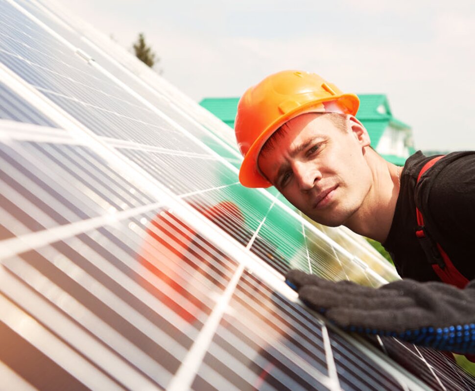 Ein Arbeiter in orangefarbener Schutzhelm und Handschuhen überprüft eine Solaranlage auf einem Dach.