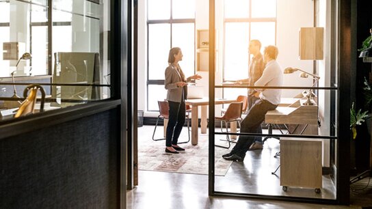 Drei Personen in einem modernen Büro diskutieren, während das Sonnenlicht durch große Fenster strömt.
