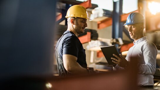 Zwei Männer in Schutzhelmen diskutieren in einem Lager, einer hält ein Tablet in der Hand.