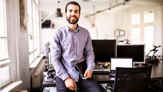 Lächelnder Mann sitzt am Schreibtisch in einem modernen Büro, umgeben von Computern und Bürotechnik.