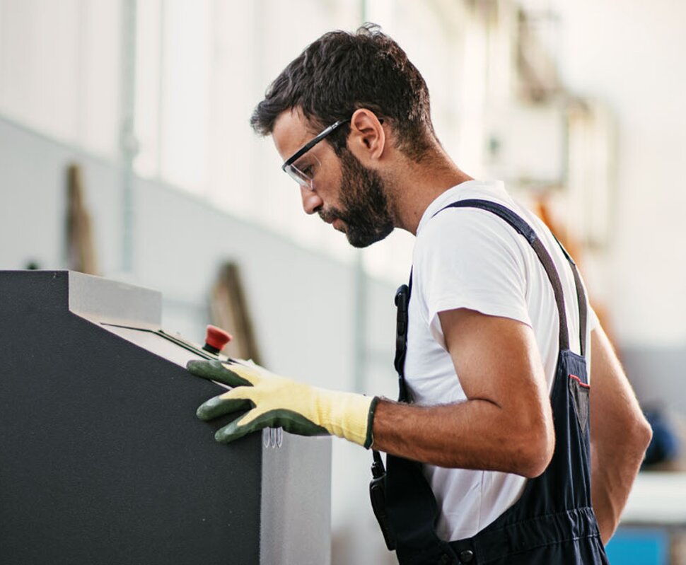 Ein Mann in Arbeitskleidung und Schutzbrille konzentriert sich auf eine Maschine in einer Industrieumgebung.