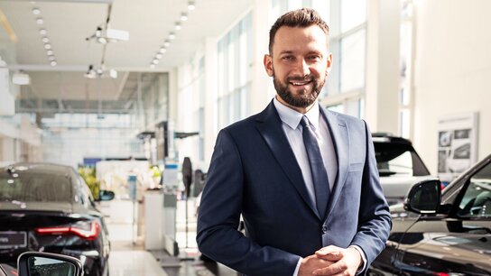 Mann im Anzug steht in einem Autohaus, umgeben von mehreren Fahrzeugen, und lächelt freundlich.