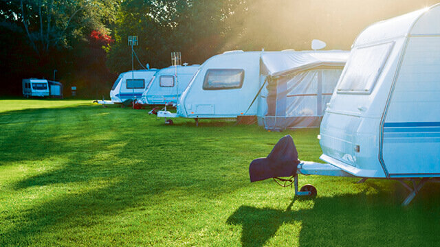 Reihen von Wohnwagen auf einer grünen Wiese, umgeben von Bäumen und Sonnenlicht, vermitteln eine entspannte Campingatmosphäre.