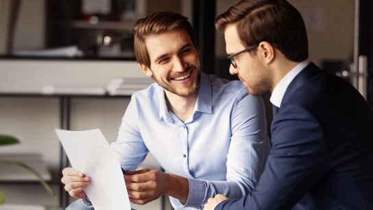 Zwei Männer im Geschäftsanzug sitzen in einem modern eingerichteten Büro und besprechen Dokumente. Sie lachen und sind freundlich.