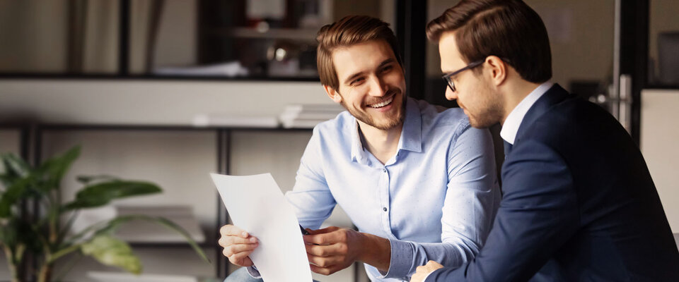 Zwei Männer im Geschäftsanzug sitzen in einem modern eingerichteten Büro und besprechen Dokumente. Sie lachen und sind freundlich.