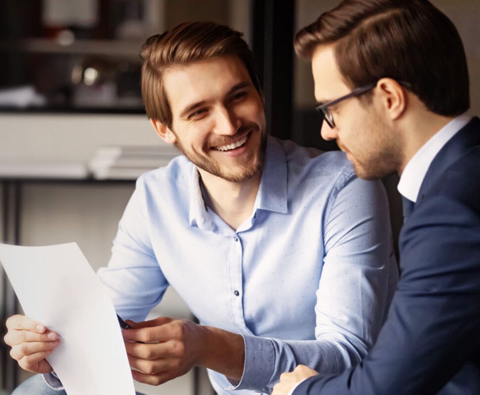 Zwei Männer im Geschäftsanzug sitzen in einem modern eingerichteten Büro und besprechen Dokumente. Sie lachen und sind freundlich.