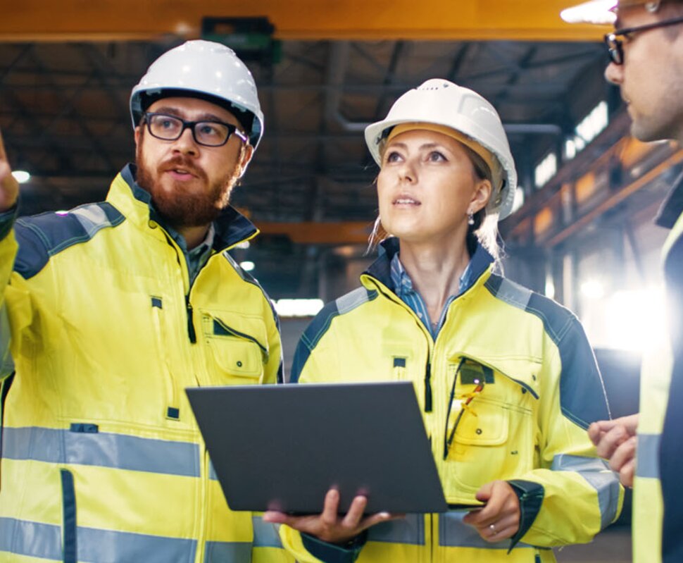 Drei Bauarbeiter in Helmen und reflektierender Kleidung besprechen Pläne mit einem Laptop in einer Lagerhalle.