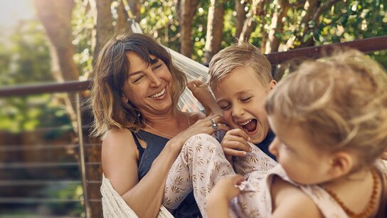 Glückliche Frau mit zwei Kindern in einer Hängematte, die gemeinsam lachen und spielen, umgeben von grüner Natur.
