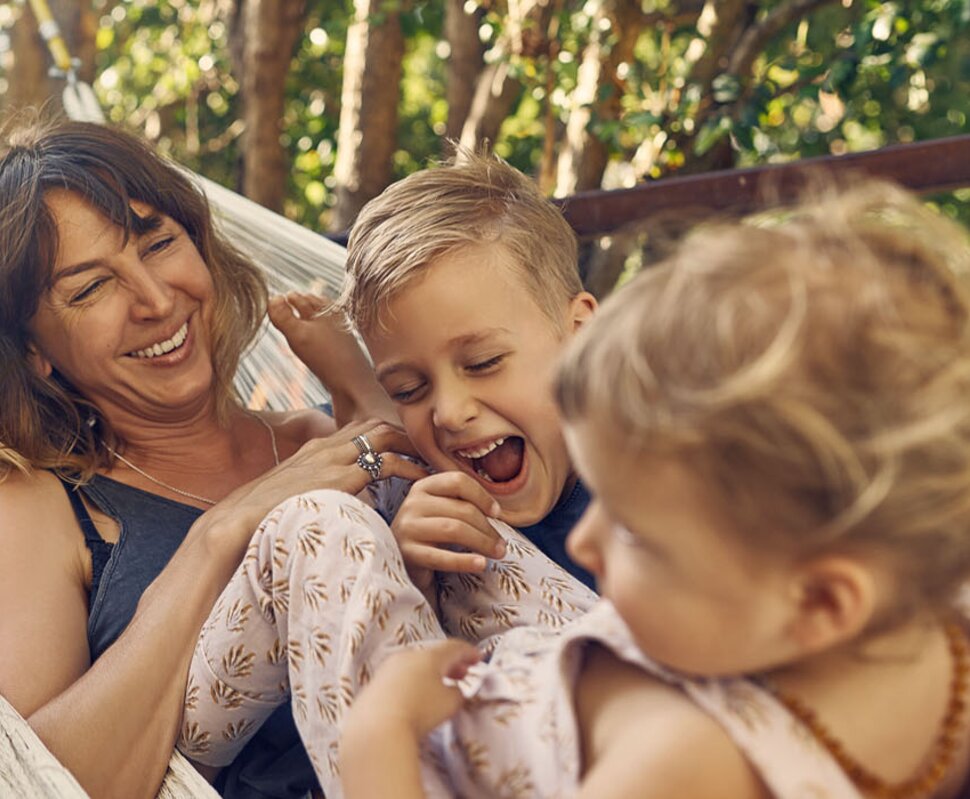 Glückliche Frau mit zwei Kindern in einer Hängematte, die gemeinsam lachen und spielen, umgeben von grüner Natur.