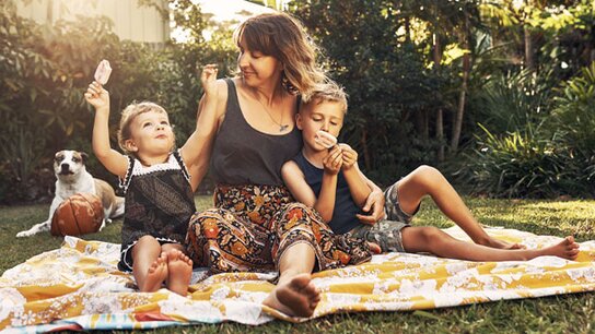 Eine Frau sitzt mit zwei Kindern auf einer Decke im Garten, alle genießen Eiscreme. Ein Hund liegt daneben.