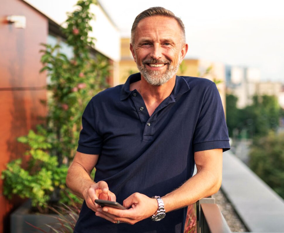Lächelnder Mann im Polohemd steht auf einem Balkon, hält ein Smartphone in der Hand, im Hintergrund Pflanzen und Stadtblick.