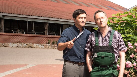 Zwei Männer stehen auf einem Hof, einer hält ein Messer, im Hintergrund ein Gebäude mit roten Dach und Blumen.