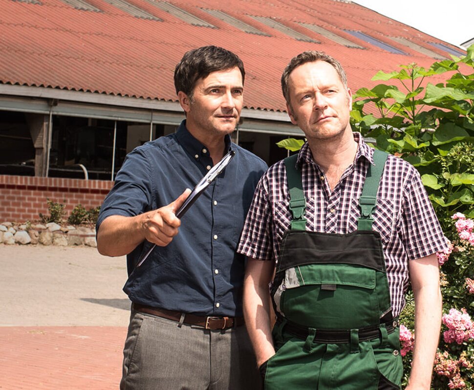 Zwei Männer stehen auf einem Hof, einer hält ein Messer, im Hintergrund ein Gebäude mit roten Dach und Blumen.