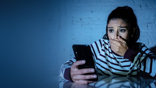 Eine junge Frau schaut besorgt auf ihr Smartphone und hält sich besorgt die Hand vor den Mund. Gedämpftes Licht von hinten.