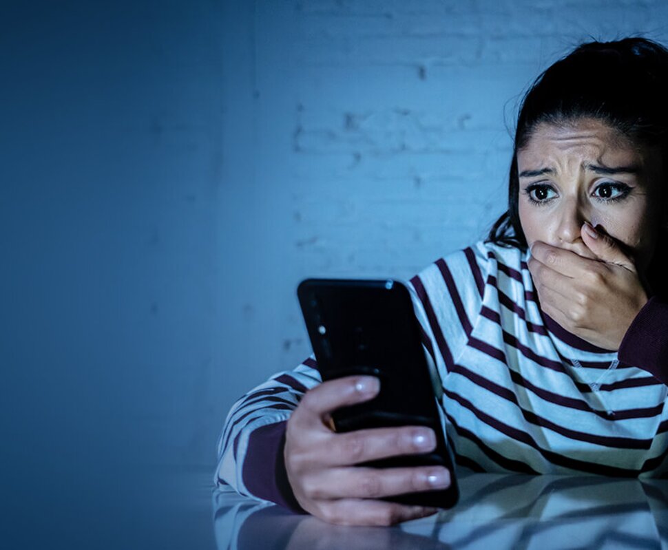 Eine junge Frau schaut besorgt auf ihr Smartphone und hält sich besorgt die Hand vor den Mund. Gedämpftes Licht von hinten.