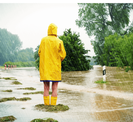 Person in gelbem Regenmantel und Gummistiefeln steht vor überfluteter Straße, während es stark regnet.