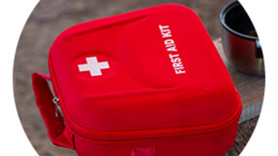 Rote Tasche mit dem Aufdruck „FIRST AID KIT“ und weißem Kreuz, auf einem Holzuntergrund platziert.