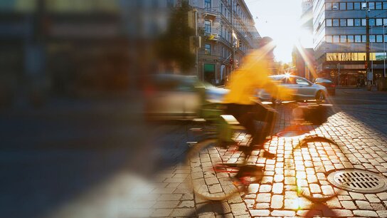 Radfahrer in gelber Jacke fährt auf einem Kopfsteinpflasterweg, Sonnenstrahlen scheinen durch die Stadt.