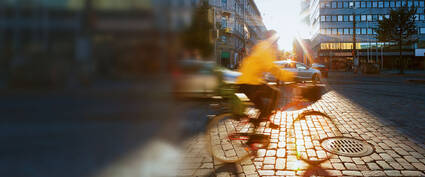 Radfahrer in gelber Jacke fährt auf einem Kopfsteinpflasterweg, Sonnenstrahlen scheinen durch die Stadt.
