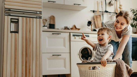 Eine Mutter schiebt einen Wäschekorb mit einem fröhlichen, lächelnden Jungen, der begeistert auf etwas zeigt, in einer hellen Küche.