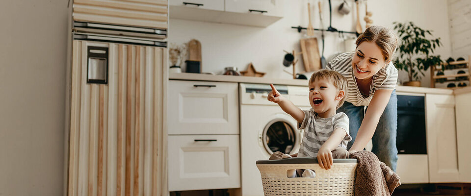 Eine Mutter schiebt einen Wäschekorb mit einem fröhlichen, lächelnden Jungen, der begeistert auf etwas zeigt, in einer hellen Küche.